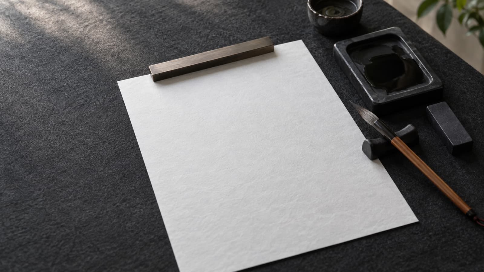 Illustration: A blank sheet of calligraphy paper on a dark felt mat, with a paperweight, an inkstone and a brush on its rest