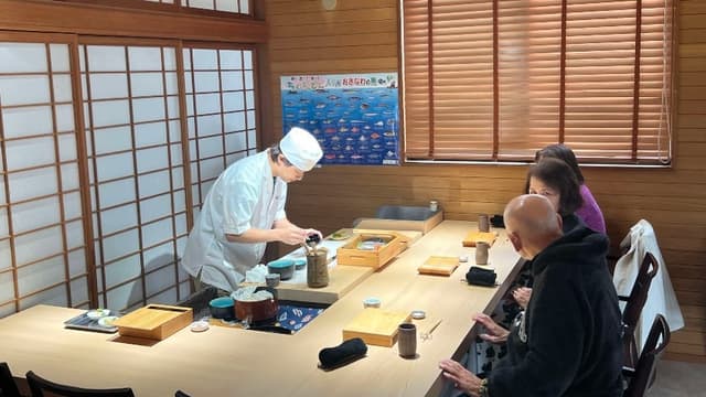 Instructor Masami Oginuma in chef's whites preparing sushi at a wooden counter for seated guests