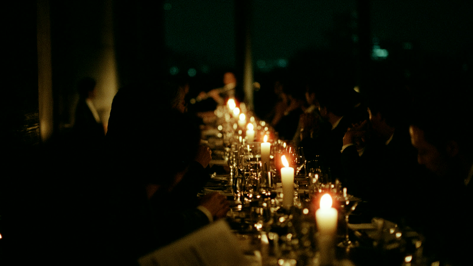 夜の東京を望む部屋で、蝋燭の灯りが一列に並ぶ長いテーブルを人々が囲んでいる（イメージ）