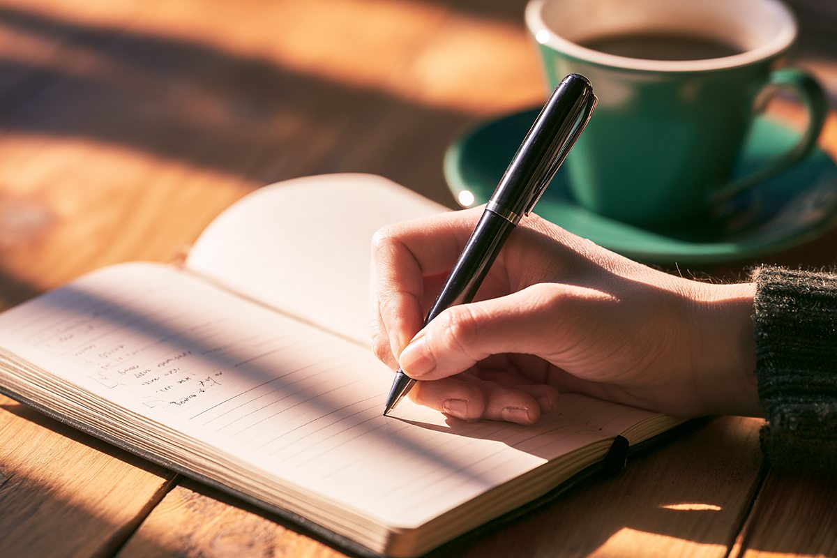 朝の光が差す木の机で、開いたノートに短い箇条書きを書きこむ手。奥にコーヒーカップ